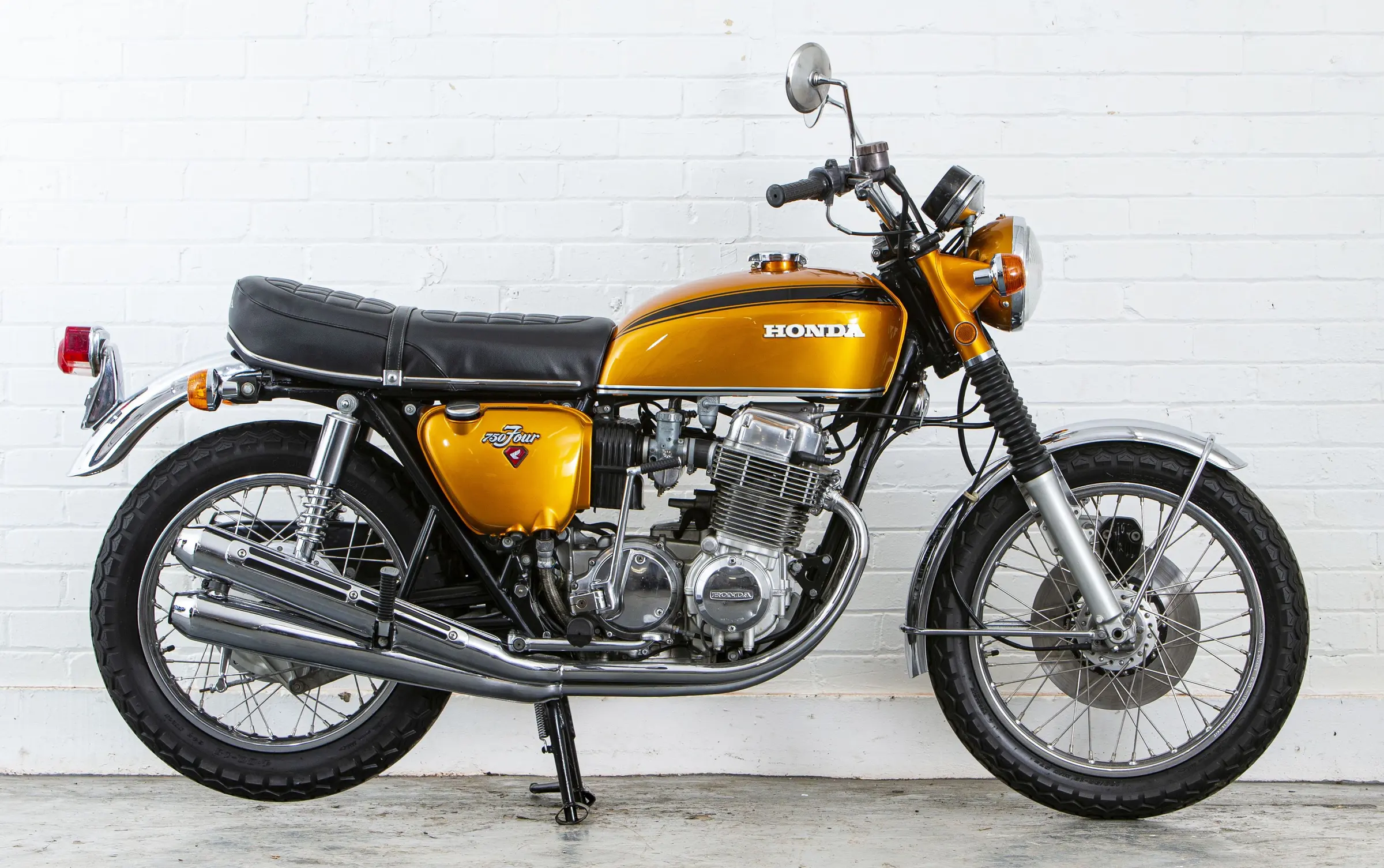 Gold Honda CB750 four-cylinder motorcycle photographed from the side against a white brick wall